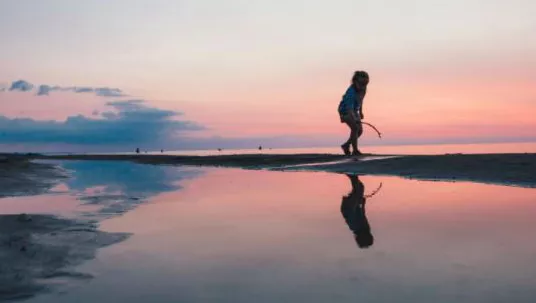 Ein Kind spielt mit einem Stock am Strand, während der Himmel in einem rosa Sonnenuntergang erstrahlt.