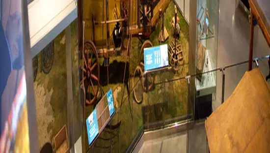 Vue de dos d'un homme se tenant debout entre deux grosses roues de chariot historique et entouré de meubles de pionniers et de photos sur les murs.