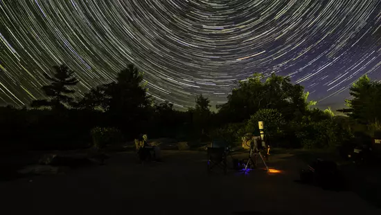 Telescopio público frente a bosques de fondo abrumados por un cielo oscuro con cientos de trayectorias de estrellas circulares paralelas brillantes