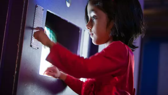 Small child standing in front of recessed computer screen pressing dots on small metal plate on the wall to her left