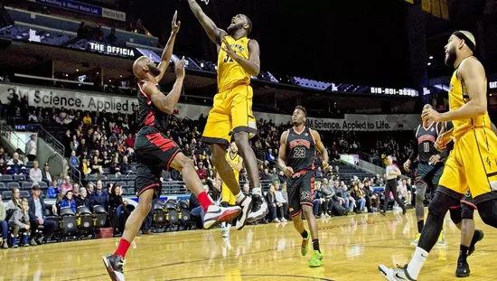 Basketball player in mid-air tossing ball into basket among players with large audience in background bleachers
