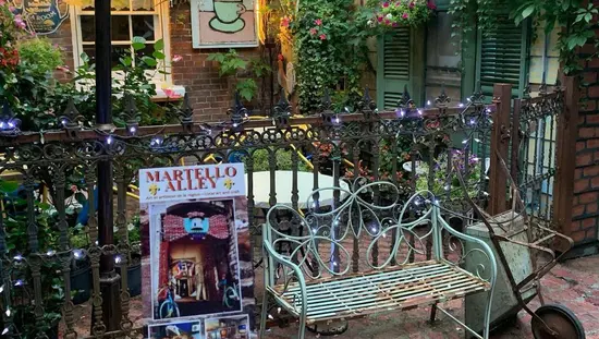 Affiche de Martello Alley à côté d'un banc vert rouillé et d'une brouette sur une ancienne promenade de brique avec un café extérieur derrière la clôture.
