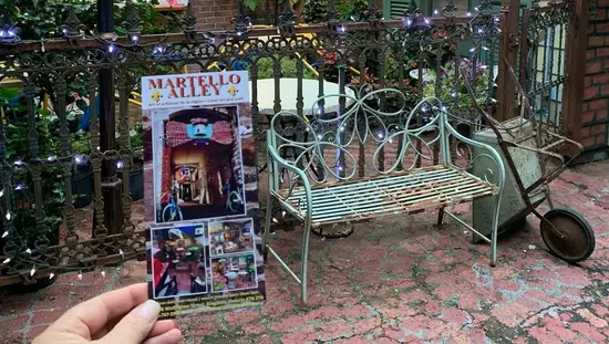 Martello Alley-Poster neben rostiger grüner Bank und Schubkarre auf altem Backsteingehweg mit Café im Freien hinter dem Zaun