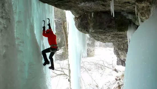 Man just beyond huge rock overhang climbing vertical wall of ice with hands gripping ice with 2 large hooks