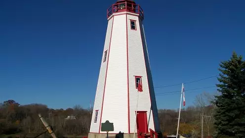 Green lawn with very tall white lighthouse with red doorway and front steps and balcony and with forest beyond