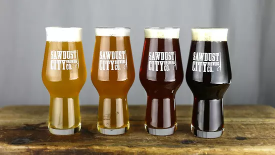 4 vasos, cada uno con un color de cerveza cada vez más oscuro, alineados en una mesa de madera antigua y cada uno con el nombre de la empresa impreso