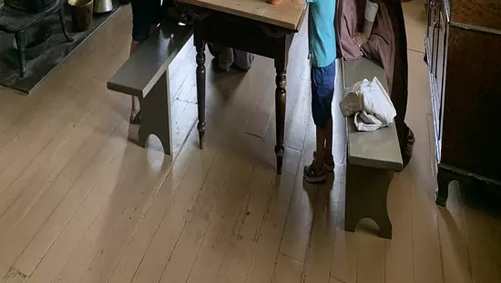 3 Frauen in langen Pionierkleidern, 2 davon am Küchentisch helfen der modernen Frau und 2 kleinen Jungen beim Brotteig kneten