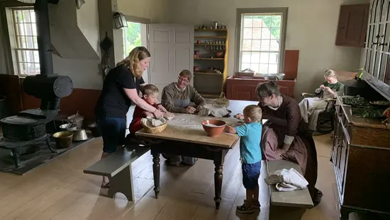 Trois femmes en robes longues de pionnières dont deux à une table de cuisine qui aident une femme moderne et deux petits garçons à pétrir de la pâte à pain.