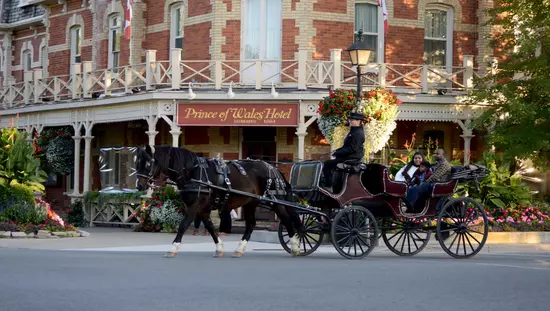 A couple with a child in an open horse-drawn carriage with a driver dressed in heritage costume, passing colourful gardens and a heritage hotel.