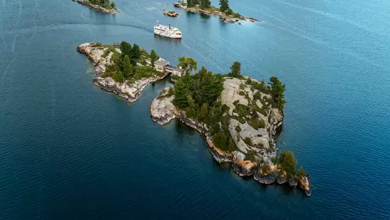 Large rivière avec deux îles, une grosse et une petite, où se trouvent des arbres et des bâtiments ainsi qu'un traversier qui effectue la traversée depuis la rive opposée au loin.