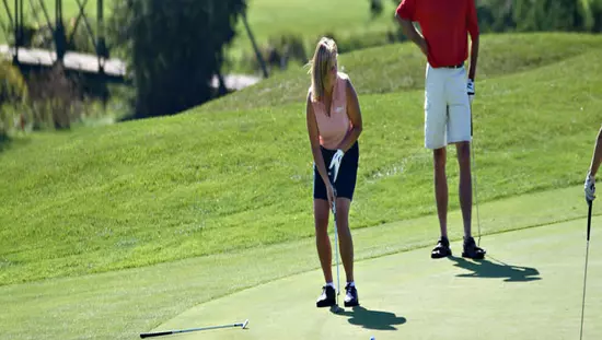 골프 코스에 있는 여성이 푸른 잔디와 상록수를 배경으로 서서 지켜보고 있는 남자와 함께 샷을 위해 골프 클럽을 줄을 서 있습니다.