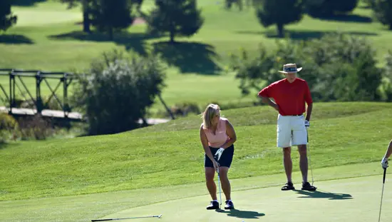 Woman on golf course lining up her golf club for a shot with man standing watching with backdrop of green grass and evergreens