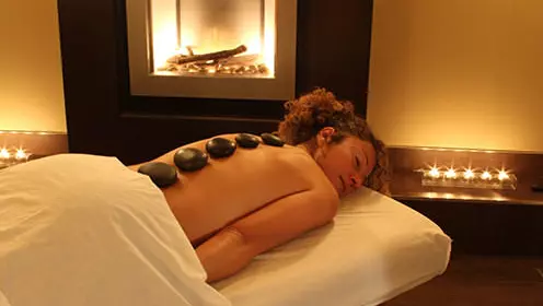 Woman lying on stomach on massage table under sheet with eyes closed and 5 large smooth brown stones spaced along her spine