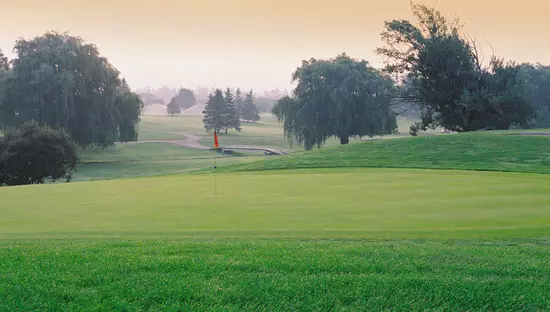 前景の穴に旗があり、木々や歩道が点在し、背景に橋がある広大なゴルフコースの芝生