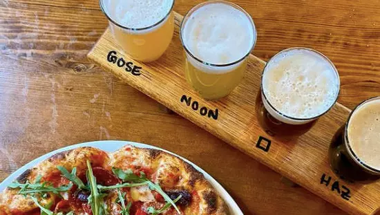 Whole pizza on plate beside board holding row of 4 glasses of beer in various colours with type of beer printed on board