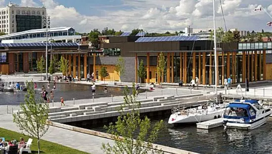 Eine Uferpromenade voller Menschen, die in einem Springbrunnenbecken faulenzen, spazieren gehen und waten, einschließlich eines Bereichs zum Anlegen von Booten und Zugang zu einem Gemeinschaftsgebäude