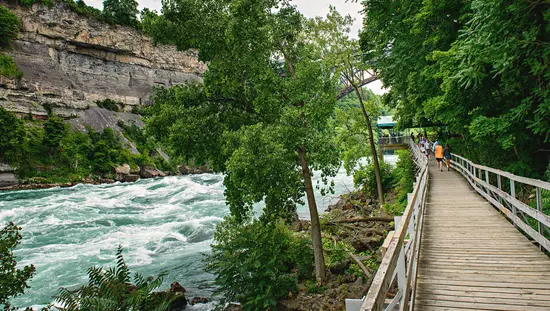 Weiße Stromschnellen fließen zwischen steilen Felswänden und Wald mit Holzsteg mit Menschen, die darauf laufen