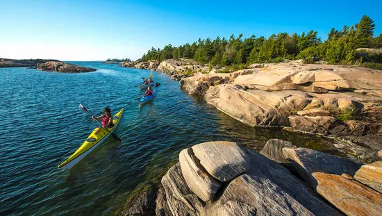 Drei Personen paddeln auf dem See in der Nähe von felsigem Land mit Bäumen.
