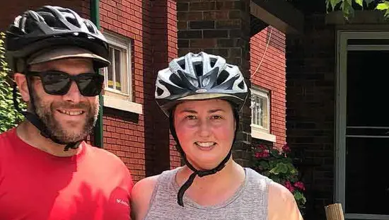 Ein Mann und eine Frau, beide mit Fahrradhelmen, posieren mit einem an einem Baum gelehnten Fahrrad
