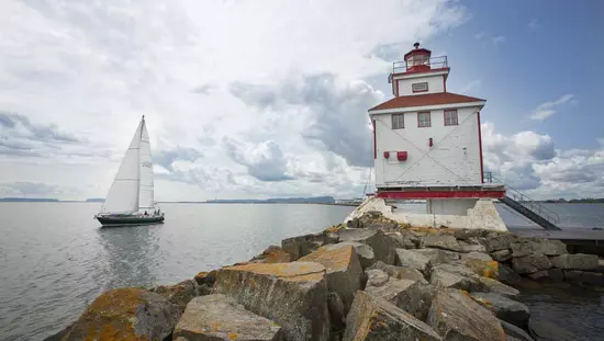 Un velero navega más allá de un faro blanco y rojo en lo alto de una costa rocosa