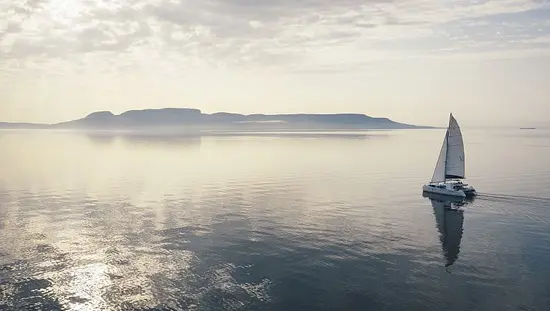 Reflections on water of a catamaran sailing towards hills in the distance as the sky is illuminated a purple gray