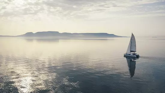 Spiegelungen auf dem Wasser eines Katamarans, der auf die Hügel in der Ferne zusegelt, während der Himmel violettgrau erleuchtet ist.