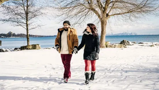 A man and a woman holding hands, walking on snow covered ground beside the lake.