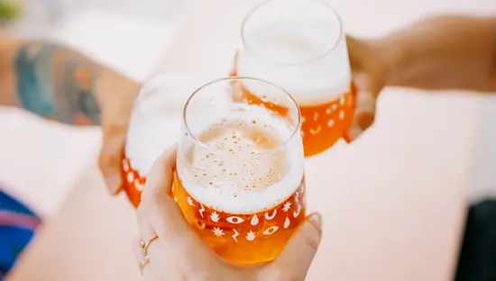 Drei Personen stoßen in entspannter Atmosphäre im Freien an einem rosa Tisch mit verspielten Mustern auf den Gläsern mit Bier an.