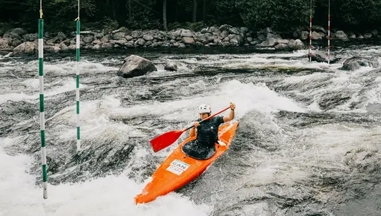 Ein Kajakfahrer befährt Wildwasserschnellen auf einem Slalomkurs und passiert dabei nummerierte Torpfosten, die über dem Fluss aufgehängt sind.