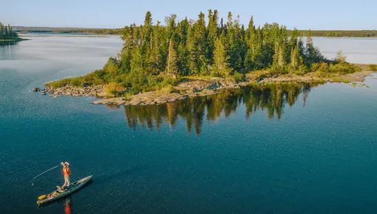  Eine Person macht Stand-Up-Paddleboarding auf einem ruhigen See und wirft unter einem klaren blauen Himmel eine Angelschnur in der Nähe einer kleinen, mit Bäumen bewachsenen Insel aus.