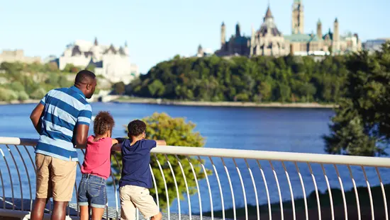 Un padre y dos niños se apoyan contra una barandilla blanca para contemplar los edificios patrimoniales al otro lado del río.