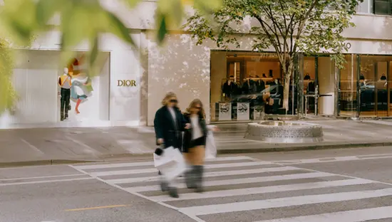 Drei Menschen gehen mit Einkaufstüten über einen Zebrastreifen vor einem Luxusgeschäftsgebäude, eingerahmt von verschwommenen grünen Blättern.