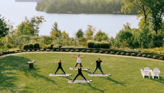 Eine kleine Gruppe praktiziert Yoga auf einer Wiese mit Blick auf einen ruhigen See, umgeben von üppigen Bäumen und fernen Inseln unter einem hellen, klaren Himmel.