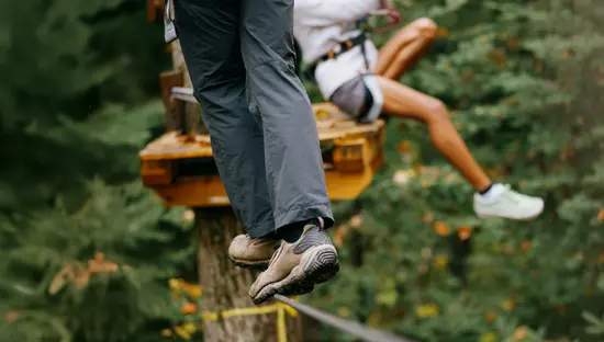 Una persona practica slackline con equipo de seguridad mientras otra, con arnés, se prepara para lanzarse en tirolesa desde una plataforma de madera en el Parque Provincial Algonquin.