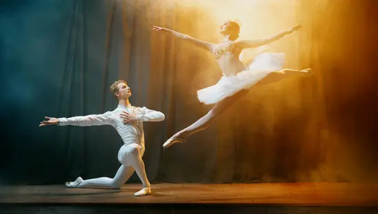 Une ballerine sautillant sur une scène peu éclairée dans un costume blanc, avec un danseur à genoux tendant les bras dans un spectacle de ballet.