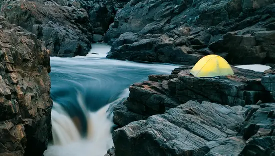 Ein leuchtendes Zelt, das auf felsigen Klippen über den Stromschnellen des Missinaibi River in der Nähe von Sault Ste. Marie in Algoma aufgestellt ist, umgeben von Wald.