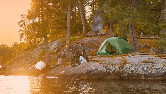 Ein Campingplatz am Opeongo Lake mit Zelt und Kanu auf einem Felsvorsprung, umrahmt von hohen Kiefern und goldenem Sonnenuntergangslicht.