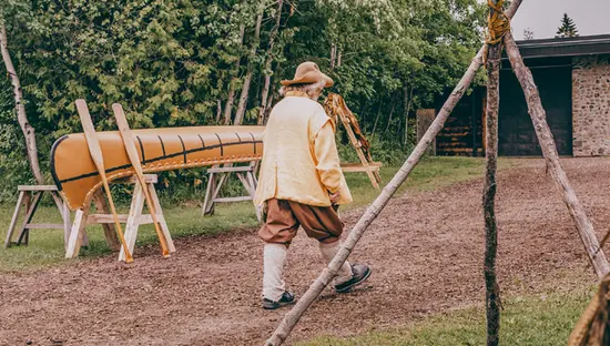Une personne en costume d'époque passant devant un canot exposé, vu de l'intérieur d'une structure autochtone avec un foyer sur l'herbe.