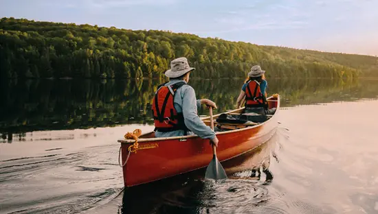 Zwei Personen paddeln in einem roten Kanu über das ruhige Süßwasser des Parry Sound, umgeben von einem üppigen Wald unter einem sanften, bewölkten Himmel.