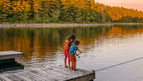 Des enfants pêchent depuis un quai au coucher du soleil, près du Crane's Lochaven Lodge, sur la rivière des Français, entourés par la forêt et l'eau.