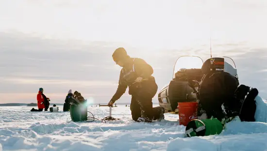 Personne pêchant sur la glace sur le lac Nipissing au lever du soleil, agenouillée à côté d'un équipement et d'une motoneige avec d'autres pêcheurs en arrière-plan.
