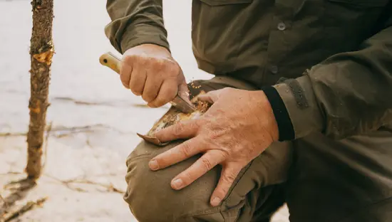 Nahaufnahme einer Person, die mit einem Messer Holz zum Anzünden neben einem See im Wabakimi Provincial Park schabt. In der Nähe liegen ein kleiner Baumstamm und Äste.