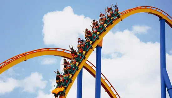 La gente se emociona subiendo a una montaña rusa, con los brazos en alto contra un cielo azul con nubes dispersas.