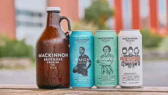A growler and three cans of Mackinnon Brothers beer—8 Man, Philomena, and Brothers House Ale—on a wooden table with red barns in the background.