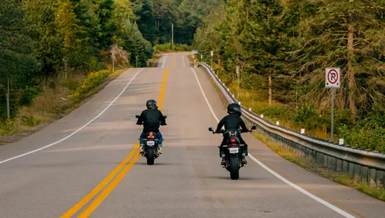 Zwei Motorradfahrer fahren an einem sonnigen Tag auf einer ruhigen Uferstraße durch die Waldlandschaft der Ontario Highlands.