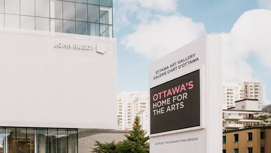 Zwei Personen gehen an einem sonnigen Tag auf den modernen Glaseingang der Ottawa Art Gallery zu.