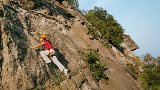 Un escalador con arnés y casco asciende por una pared rocosa empinada en el Área de Conservación de Rattlesnake Point en un día soleado, con cuerdas aseguradas arriba.