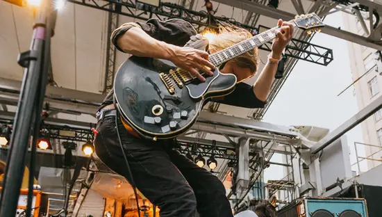  Banda de rock tocando en la plaza Yonge-Dundas en el centro de Toronto, con un guitarrista saltando junto a sus compañeros de banda y edificios altos detrás.