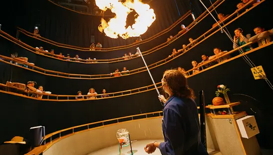 Un educador científico enciende un experimento con fuego en el escenario mientras visitantes curiosos observan desde asientos escalonados en Science North en Sudbury, Ontario.