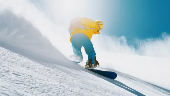 明るい色のギアを身に着けたスノーボーダーが、晴れた青空の下、太陽が降り注ぐ斜面で新雪のスノーボードを楽しんでいます。